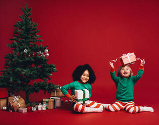 Zwei Kinder sitzen mit ihren Weihnachtsgeschenken neben einem Weihnachtsbaum. Kleines Mädchen wirft ihr Geschenk in der Luft sitzt mit einem anderen Mädchen neben einem geschmückten Weihnachtsbaum und Geschenk-Boxen. - JLPSF20439
