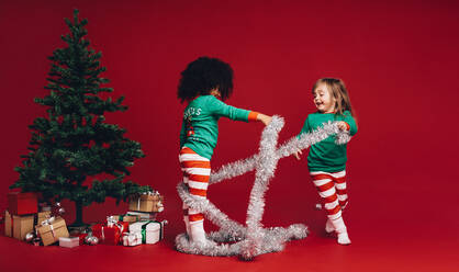 Kinder spielen mit dekorativen Band in der Nähe eines Weihnachtsbaums. Multi ethnische Kinder spielen neben einem Weihnachtsbaum und Geschenke. - JLPSF20440