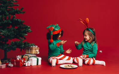Kleine Mädchen sitzen neben einem Weihnachtsbaum und genießen Kekse und Milch. Zwei glückliche Kinder in weihnachtliche Kleidung gekleidet sitzen auf dem Boden neben einem Weihnachtsbaum und Geschenk-Boxen. - JLPSF20442