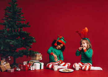 Zwei kleine Mädchen in weihnachtlichen Kleidern sitzen neben einem Weihnachtsbaum. Glückliche Kinder sitzen neben einem Weihnachtsbaum und genießen Kekse mit Milch. - JLPSF20443