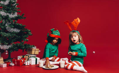 Kinder sitzen neben einem geschmückten Weihnachtsbaum in schicken Kostümen. Kinder essen Kekse mit Milch und sitzen neben einem Weihnachtsbaum und Geschenkboxen. - JLPSF20444