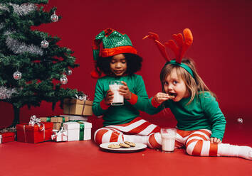 Zwei Kinder sitzen neben einem Weihnachtsbaum und essen Kekse mit Milch. Kleine Mädchen in schicken Weihnachtskleidern sitzen neben einem Weihnachtsbaum mit Geschenken. - JLPSF20446