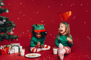 Kleine Mädchen genießen die künstlichen Schneeflocken, die neben einem Weihnachtsbaum sitzen. Kinder in schicken Weihnachtskleidern sitzen neben einem Weihnachtsbaum und essen Kekse mit Milch. - JLPSF20447