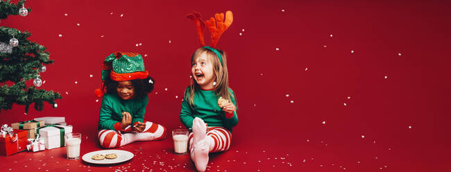 Kleine Mädchen genießen die künstlichen Schneeflocken, die neben einem Weihnachtsbaum sitzen. Kinder in schicken Weihnachtskleidern sitzen neben einem Weihnachtsbaum und essen Kekse mit Milch. - JLPSF20448