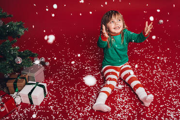 Kleines Mädchen sitzt auf dem Boden und genießt den künstlichen Schneefall. Ein Kind hat Spaß beim Spielen mit Schneeflocken, während es neben einem geschmückten Weihnachtsbaum sitzt. - JLPSF20450