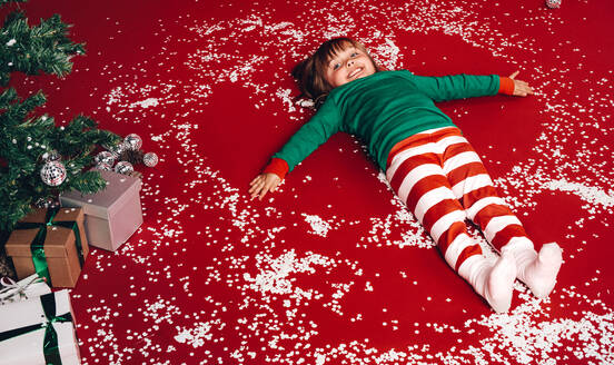 Glückliches kleines Mädchen auf dem Boden liegend neben einem Weihnachtsbaum. Kind hat Spaß beim Spielen mit Schneeflocken auf dem Boden liegend neben einem geschmückten Weihnachtsbaum. - JLPSF20451