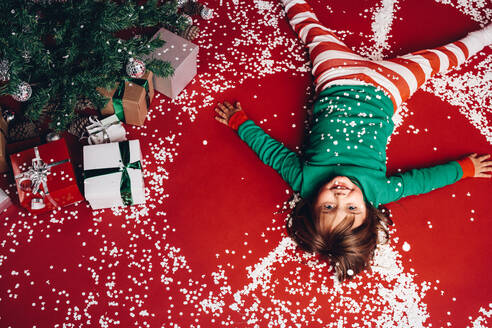 Draufsicht auf ein Kind, das auf dem Boden liegt, mit künstlichen Schneeflocken überall. Kleines Mädchen hat Spaß, wenn es neben einem Weihnachtsbaum auf dem Boden liegt - JLPSF20452