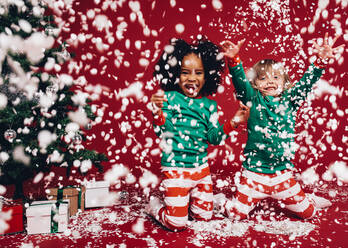 Zwei kleine Mädchen in weihnachtlichen Kostümen spielen mit künstlichen Schneeflocken. Kinder haben Spaß an einem künstlichen Schneefall neben einem Weihnachtsbaum. - JLPSF20455