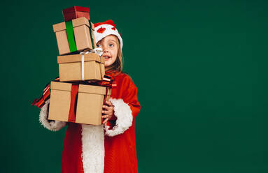 Kleines Mädchen im Nikolauskleid, das einen Stapel Geschenkkartons in der Hand hält. Kind als Weihnachtsmann verkleidet, das Geschenke trägt. - JLPSF20464