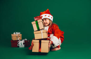 Kleines Mädchen im Weihnachtsmannkleid hebt einen Stapel Geschenkkartons. Kind im Weihnachtsmannkostüm spielt mit Geschenkkartons. - JLPSF20467