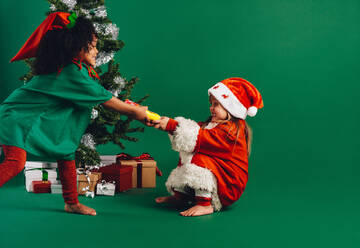 Zwei kleine Mädchen im Weihnachtskostüm ziehen ein Spielzeug. Kinder streiten sich um ein Spielzeug, das neben einem geschmückten Weihnachtsbaum steht. - JLPSF20471