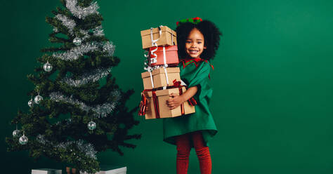 Kind hält ihre Weihnachtsgeschenkboxen neben einem geschmückten Weihnachtsbaum stehend. Glückliches afroamerikanisches Mädchen, das einen Stapel von Geschenkboxen trägt. - JLPSF20474