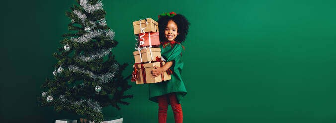 Kind hält ihre Weihnachtsgeschenkboxen neben einem geschmückten Weihnachtsbaum stehend. Glückliches afroamerikanisches Mädchen, das einen Stapel von Geschenkboxen trägt. - JLPSF20475