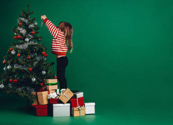 Kleines Mädchen schmückt einen Weihnachtsbaum auf Geschenkkartons stehend. Kind streckt seine Arme hoch, um einen Weihnachtsbaum zu schmücken. - JLPSF20477