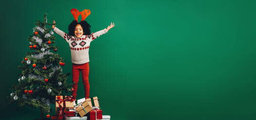Glückliches kleines Mädchen, das mit ausgestreckten Armen auf Geschenkkartons neben einem geschmückten Weihnachtsbaum steht. Kind aufgeregt nach dem Schmücken eines Weihnachtsbaums. - JLPSF20480