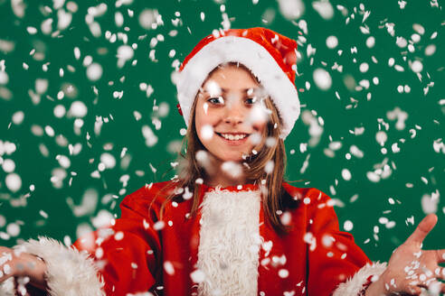 Lächelndes Mädchen im Weihnachtsmannkleid vor grünem Hintergrund mit künstlichen Schneeflocken, die rundherum fallen. Porträt eines lächelnden Mädchens mit offenen Armen, das sich über künstlichen Schneefall freut. - JLPSF20487