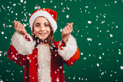 Porträt eines lächelnden Mädchens, das sich über künstlichen Schneefall freut. Fröhliches Mädchen als Weihnachtsmann verkleidet vor grünem Hintergrund mit künstlichen Schneeflocken, die rundherum fallen. - JLPSF20488