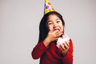 Porträt eines kleinen Mädchens, das Zuckerwatte isst. Ein Kind, das eine Party-Kegelmütze trägt, genießt Zuckerwatte. - JLPSF20500