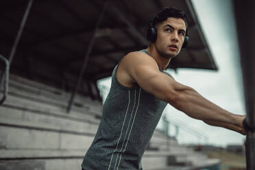 Fit young man in sportswear wearing headphone to listen music during outdoor training. Sportsman taking break from training and listening music at the stadium. - JLPSF20511