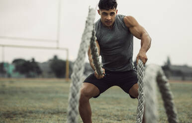 Muscular young man working out with battling ropes. Fit young male athlete doing battle rope workout outdoors on a field. - JLPSF20514