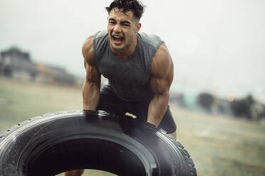 Kräftiger junger Sportler bei einer Reifenwendeübung im Regen. Muskulöser Mann beim Crosstraining im Freien auf einem Feld. - JLPSF20519