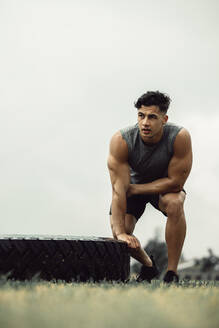 Ein fitter junger Mann, der im Freien einen Reifensprung macht. Ein muskulöser Athlet, der eine Pause vom Cross-Training auf einem Feld macht. - JLPSF20521