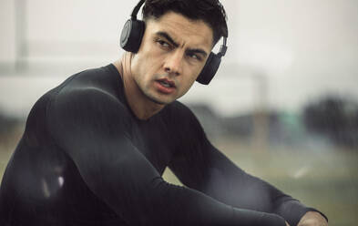 Fit male runner relaxing after workout in rainy morning. Muscular young man with headphones on listening to music while taking a break from training. - JLPSF20525