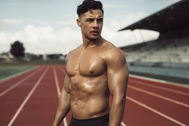 Runner standing on a running track and looking away. Bare chested muscular athlete relaxing after a run standing on the track. - JLPSF20531