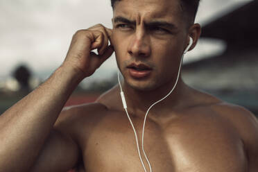 Nahaufnahme eines fitten jungen Mannes, der Kopfhörer trägt, um Musik zu hören. Ein muskulöser männlicher Sportler im Freien, der eine Trainingspause einlegt. - JLPSF20538