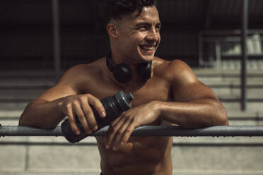 Lächelnder junger Sportler mit Wasserflasche am Geländer des Sportstadions lehnend. Hemdloser junger Mann, der sich nach dem Training ausruht. - JLPSF20545