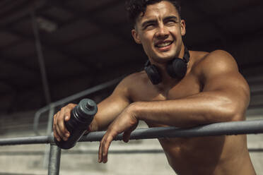 Muskulöser Mann mit Wasserflasche, der sich auf das Geländer des Stadions stützt, wegschaut und lächelt. Mann, der sich nach dem Training im Stadion ausruht. - JLPSF20546