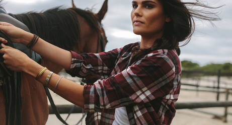 Junges Cowgirl, das einem braunen Pferd den Sattel anlegt, bevor es reitet. Eine Reiterin macht ihr Pferd für einen Ausritt bereit. - JLPSF20549