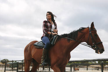 Junge Frau auf ihrem Pferd sitzend und wegschauend, weiblicher Jockey reitet auf ihrem Pferd im Korral. - JLPSF20553