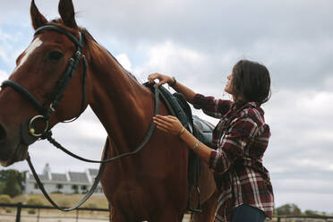 Junge Frau bereitet ihr Pferd für einen Ausritt vor. Junge Frau reitet mit ihrem Pferd und legt den Sattel auf den Rücken im Stall. - JLPSF20554