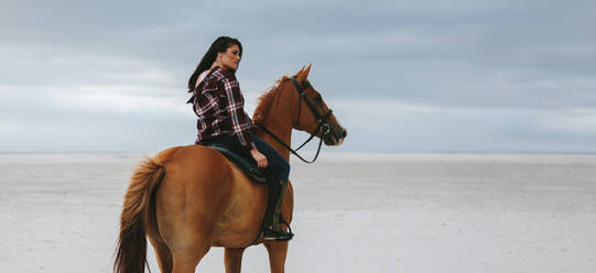 Portrait of young cowgirl riding on horse at beach. Woman horse riding at the sea shore and looking away. - JLPSF20559