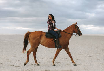 Frau reitet am Strand bei Sonnenuntergang und blickt zurück. Schöne Frau reitet auf einem braunen Pferd am Abend. - JLPSF20561