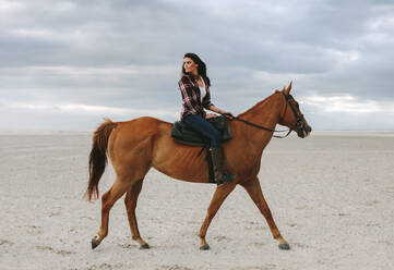 Woman horse ride on beach at sunset and glancing back. Beautiful female riding a brown horse in evening. - JLPSF20561