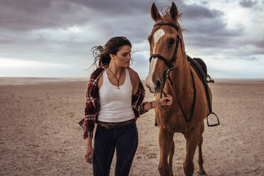 Porträt einer Frau, die mit einem Pferd am Strand spazieren geht. Eine Reiterin geht mit ihrem Hengst am Abend spazieren. - JLPSF20575