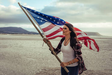 Attractive young woman holding an American Flag while standing on the beach. Young woman in casuals with the USA flag. - JLPSF20577