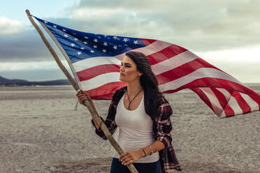 Schöne kaukasische Frau hält eine amerikanische Flagge, während sie am Meer steht. Junge Frau mit der USA-Flagge am Strand. - JLPSF20578