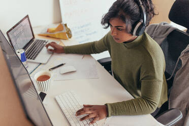 Eine Programmiererin programmiert an einem Desktop-Computer und einem Laptop an ihrem Schreibtisch im Büro. Junge Frau in Freizeitkleidung mit Kopfhörern arbeitet an einem Laptop und einem Desktop-Computer. - JLPSF20587