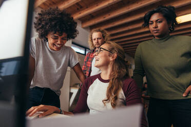 Multi-ethnic startup business team working together in office. Woman sitting at her desk laughing with team standing by. Business team having casual talk while working. - JLPSF20604