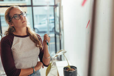 Junge Frau, die im Büro steht und auf die Präsentationstafel schaut, denkt nach. Weibliche Computerprogrammiererin, die über einen neuen Projektplan bei einem Tech-Startup nachdenkt. - JLPSF20610