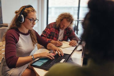 Computerprogrammierer arbeiten in einem Coworking Space in einem Startup-Büro. Eine Frau mit Kopfhörern programmiert am Laptop, während ihre Kollegen um den Tisch herum arbeiten. - JLPSF20620