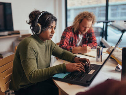 Computerprogrammierer mit Kopfhörern, die an einem Laptop programmieren, mit Kollegen, die in einem Startup-Büro arbeiten. Junge Berufstätige arbeiten in einem Büro eines Tech-Startups. - JLPSF20621