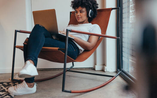 Junge afrikanische Frau sitzt entspannt auf einem Stuhl, trägt Kopfhörer und arbeitet an einem Laptop. Software-Programmiererin arbeitet in einem Startup-Unternehmen. - JLPSF20627