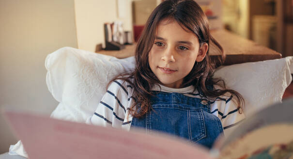 Young girl sitting at home reading a book. Cute caucasian girl student engrossed in reading story book indoors. - JLPSF20631