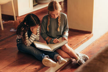 Mädchen liest Märchenbuch für ihre kleine Schwester. Zwei Mädchen sitzen auf dem Boden und lesen ein Buch zu Hause. - JLPSF20633