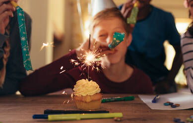 Cupcake on table with sparkler and children blowing party whistles. Kids celebrating at birthday party. - JLPSF20634