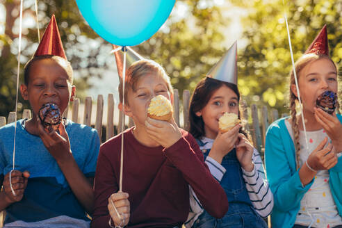 Eine Gruppe von Kindern mit Partyhüten, die Luftballons halten und Muffins auf einer Party essen. Kleine Mädchen und Jungen feiern zusammen. - JLPSF20635